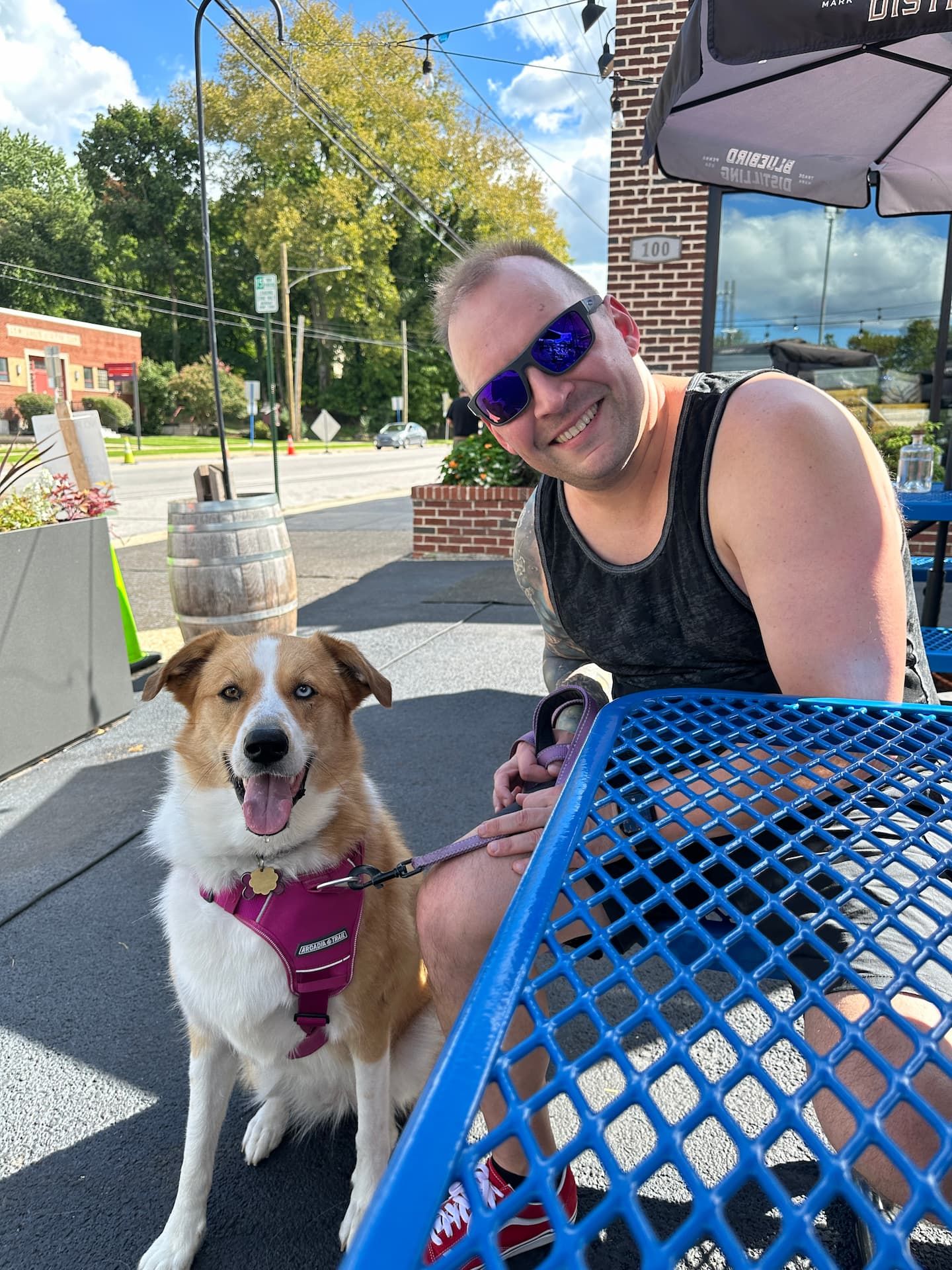 Tess and I sitting at an outside table in front of a brick building. Tess is a tan and white dog smiling and I'm wearing sunglasses smiling.