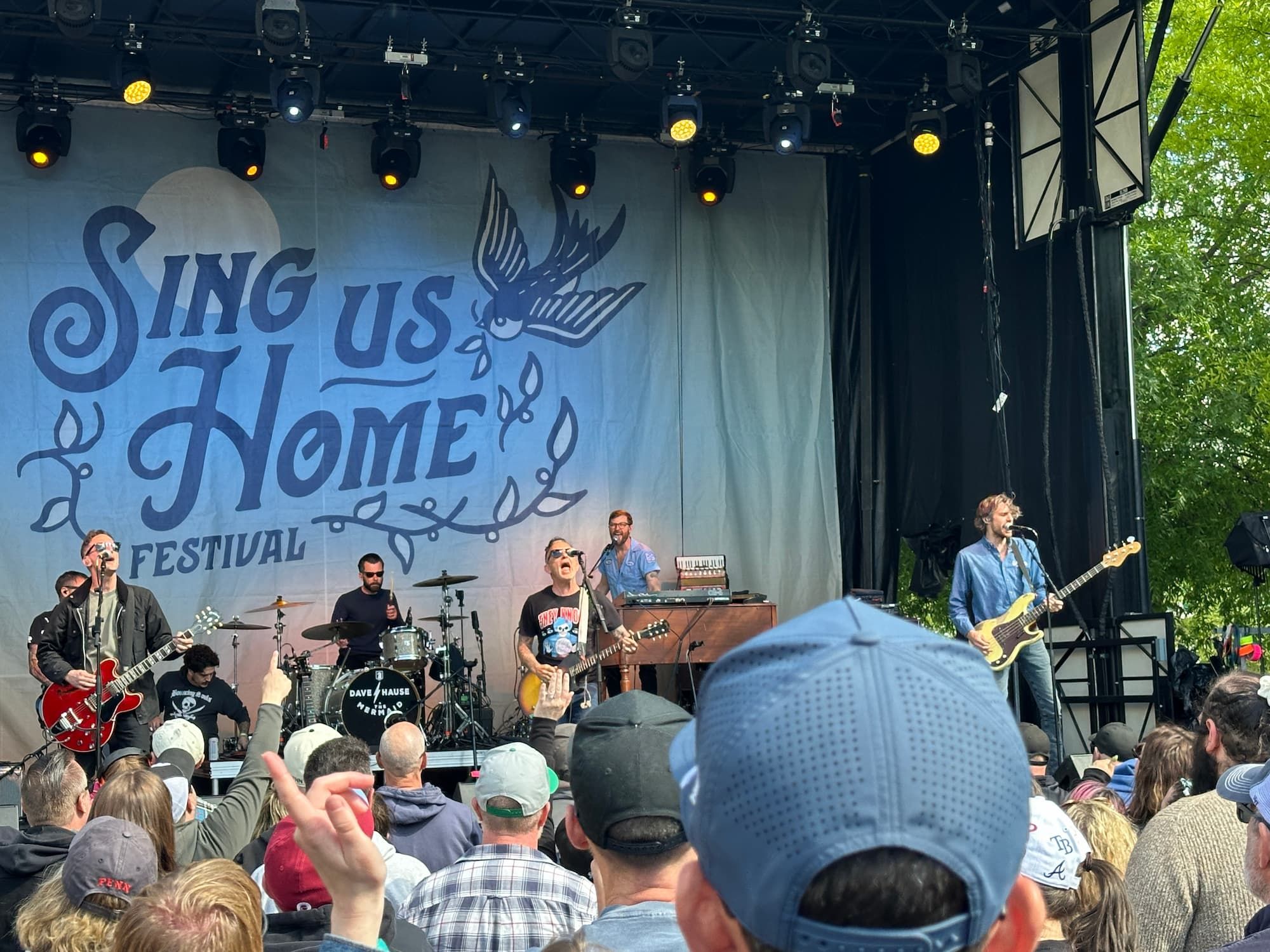 Dave Hause & the Mermaid playing live on stage with a Sing Us Home poster behind them
