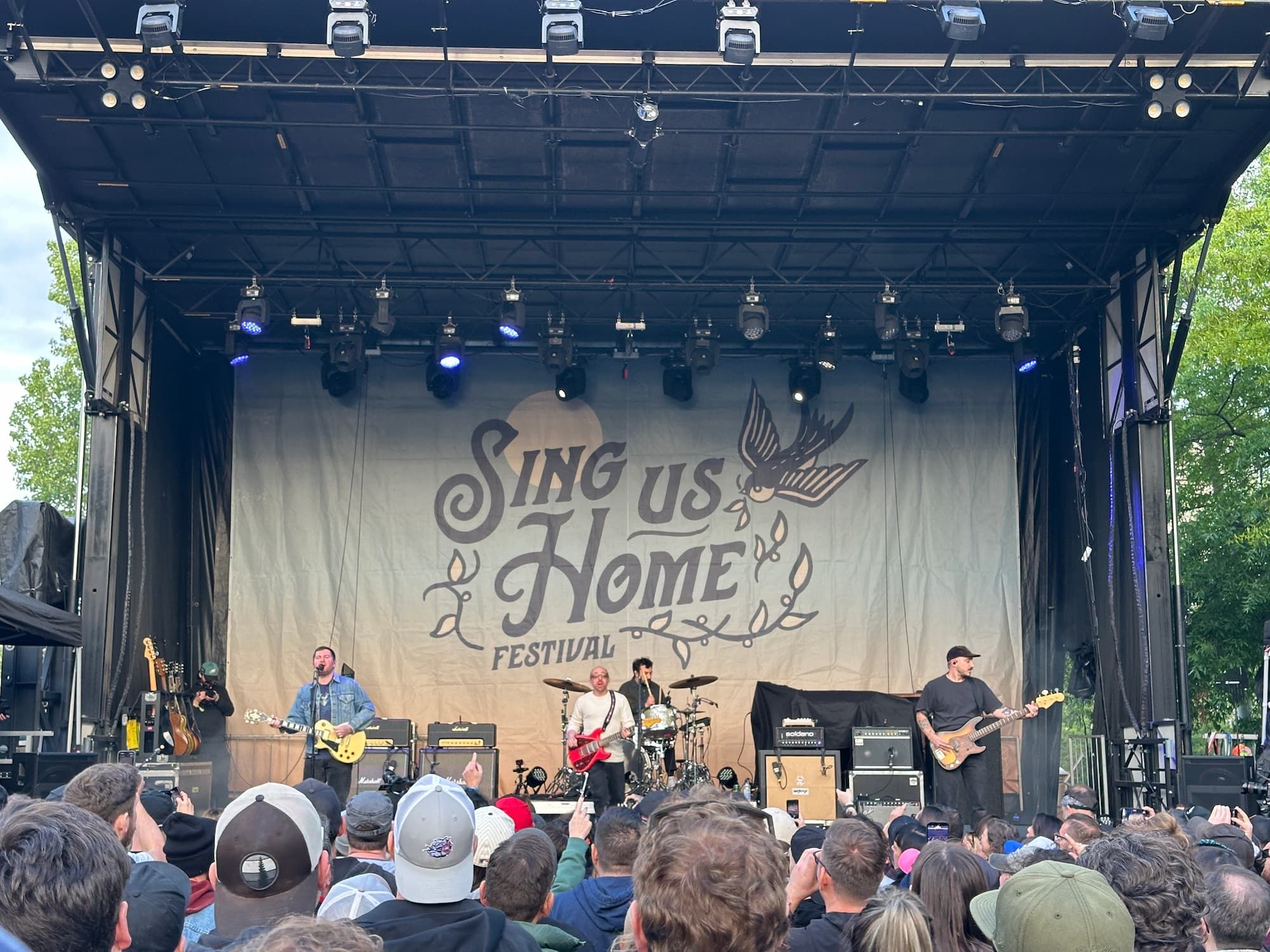 The Menzingers playing live on stage with a Sing Us Home poster behind them